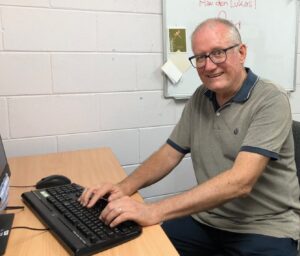 Romero Centre volunteer, Joe, sits working at his computer while smiling at the camera.